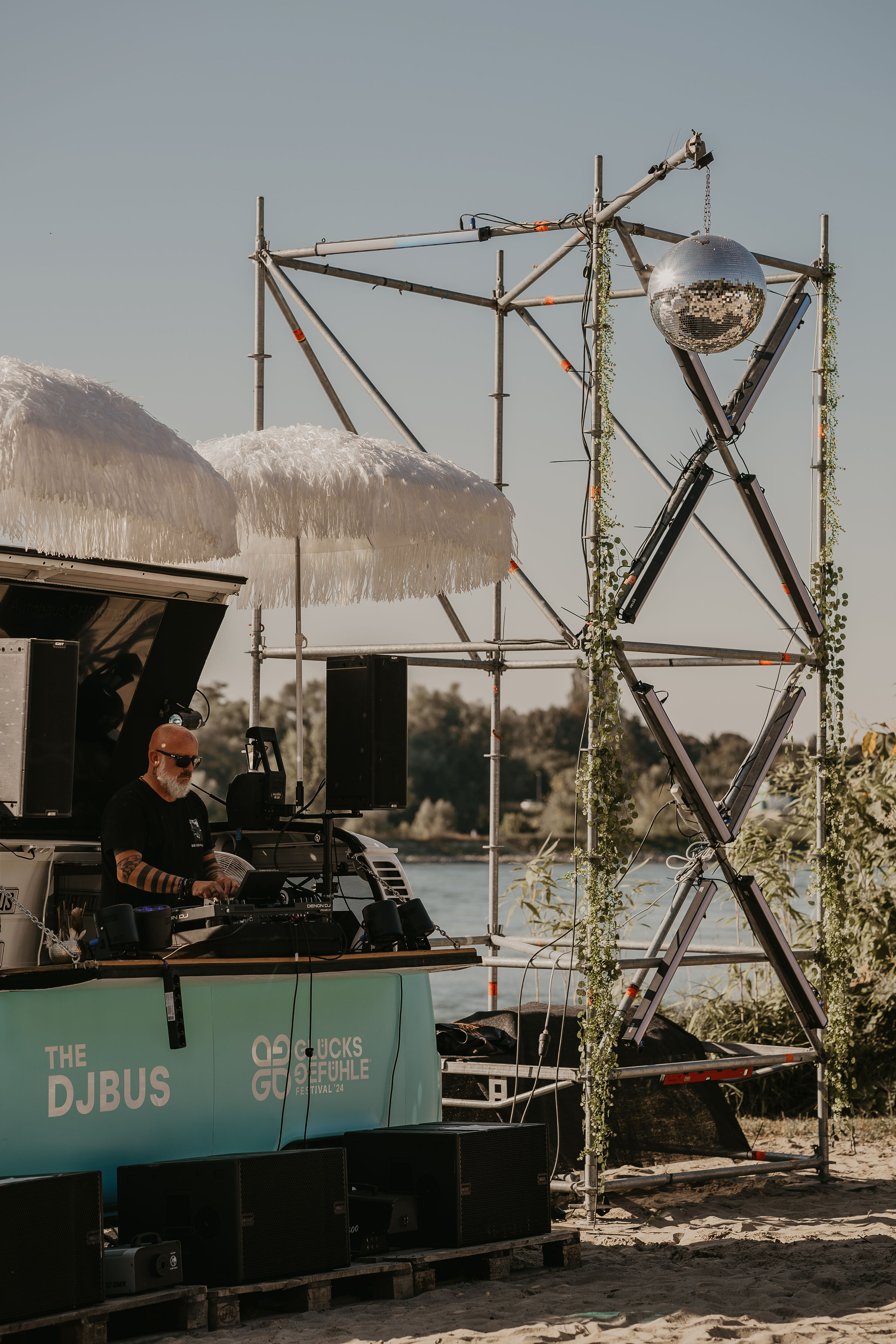 DJ an einem mobilen DJ-Bus am Rheinufer mit Discokugel und Frangipani-Sonnenschirm