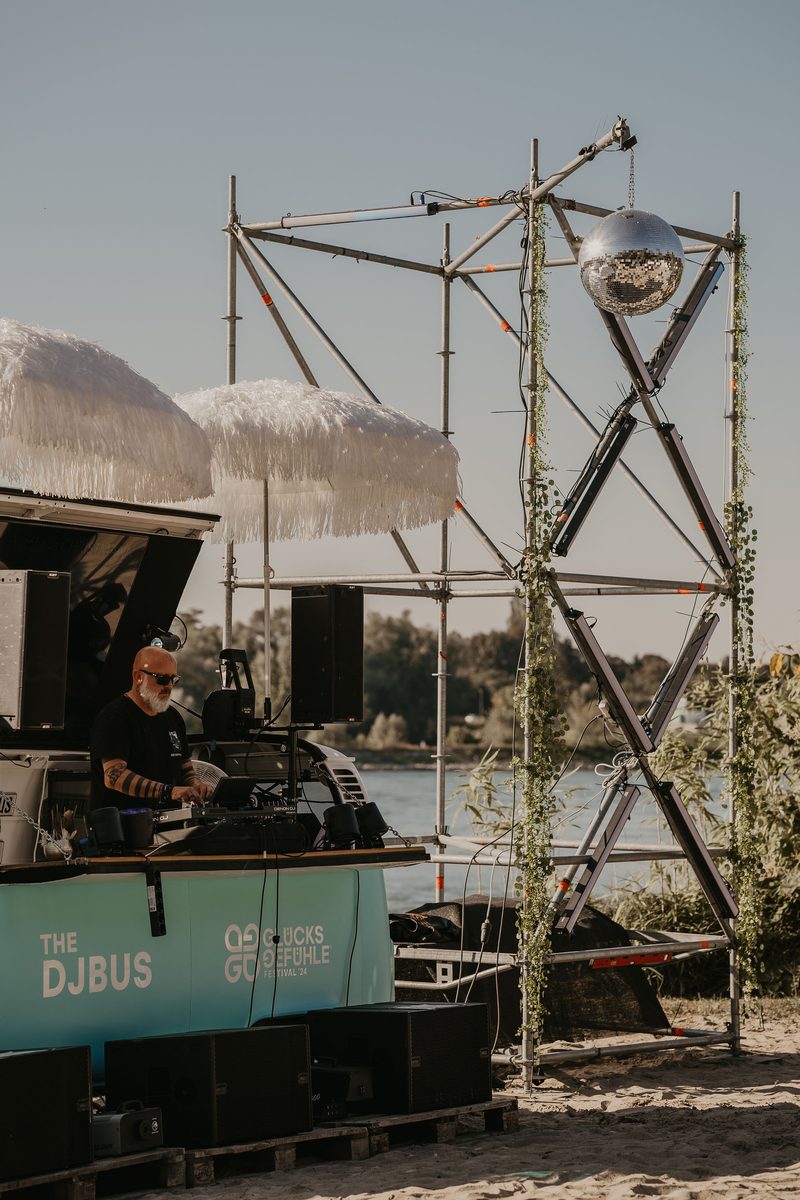 DJ an einem mobilen DJ-Bus am Rheinufer mit Discokugel und Frangipani-Sonnenschirm