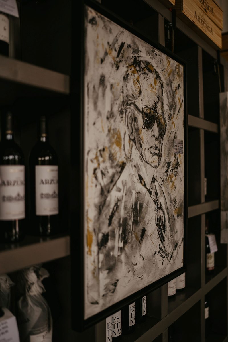 Gerahmtes Porträtgemälde einer Person mit Sonnenbrille zwischen Weinregalen bei der House Art Ausstellung